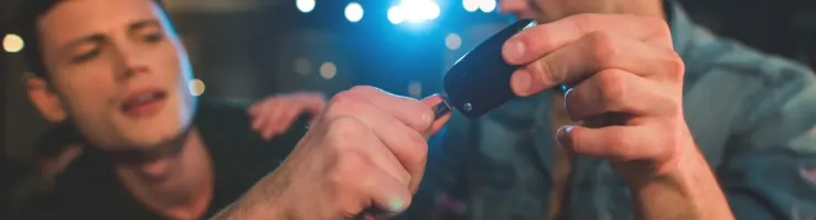 Deux jeunes hommes dans un lieu sombre, l'un tendant une clé de voiture à l'autre, avec des lumières floues en arrière-plan.