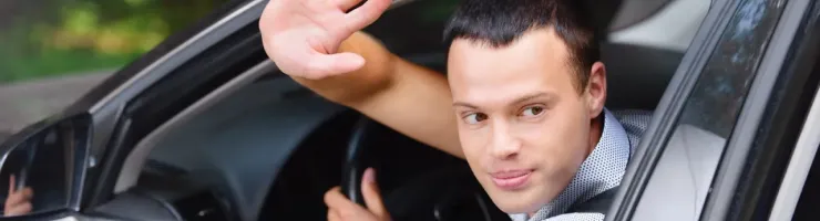 Un homme souriant dans une voiture noire lève la main pour dire au revoir ou saluer, regardant hors de la fenêtre ouverte.