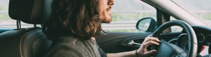 Un jeune homme aux longs cheveux et à la barbe conduit une voiture, le regard fixé devant lui, montrant son profil.