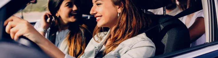 Une jeune femme souriante conduit une voiture, avec deux amies assises à l'arrière, profitant du voyage sous le soleil.