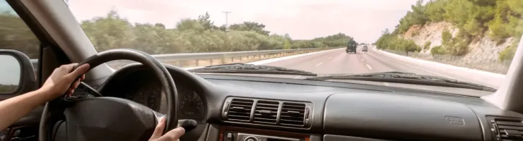 Vue depuis l'intérieur d'une voiture, les mains d'une personne tenant le volant, roulant sur une autoroute bordée d'arbres.