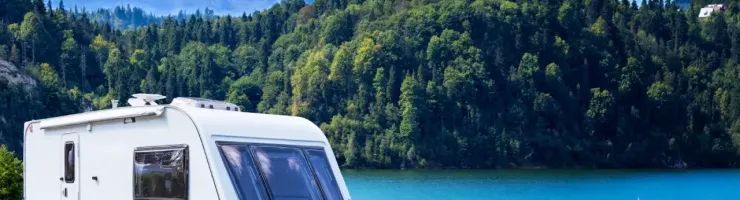 Une caravane blanche est stationnée au bord d'un lac, avec des montagnes et une forêt en arrière-plan sous un ciel bleu.
