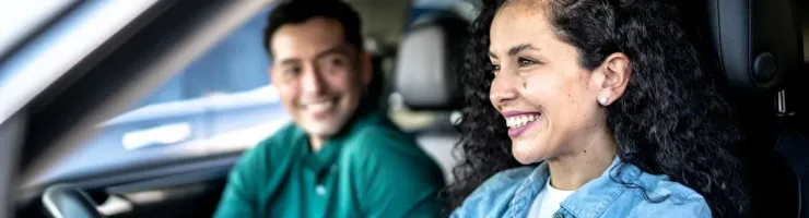 Une jeune femme souriante conduit une voiture, avec un homme souriant sur le siège passager.