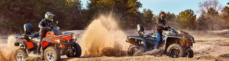 Deux personnes en quads soulèvent de la poussière en roulant sur un terrain sablonneux, avec des arbres en arrière-plan.