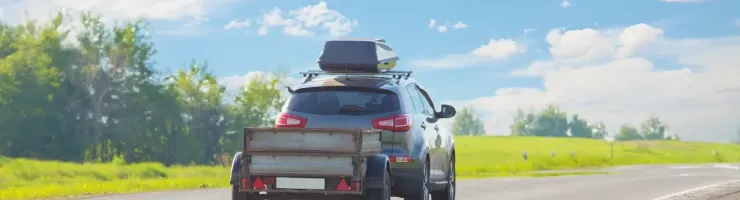 Un SUV gris avec un coffre de toit et une remorque roule sur une route de campagne sous un ciel bleu nuageux.