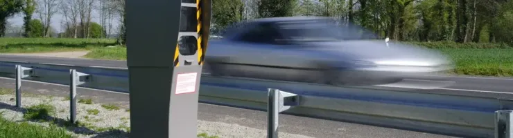 Un radar fixe est installé au bord d'une route, avec une voiture en mouvement rapide et floue en arrière-plan.