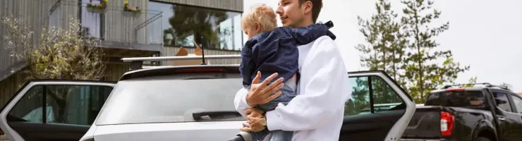 Un homme porte un jeune enfant près d'une voiture familiale blanche avec les portes ouvertes.