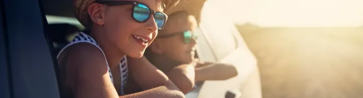 Deux enfants souriants portent des chapeaux de paille et des lunettes de soleil, regardant par la fenêtre d'une voiture.