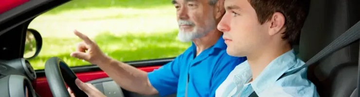 Un jeune homme conduit une voiture rouge, un homme plus âgé à ses côtés pointe du doigt, enseignant la conduite.