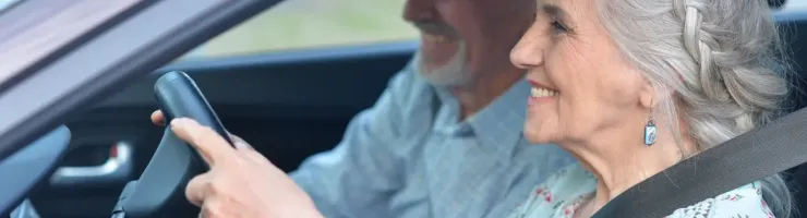 Un couple de personnes âgées souriantes est assis dans une voiture, la femme tient le volant.