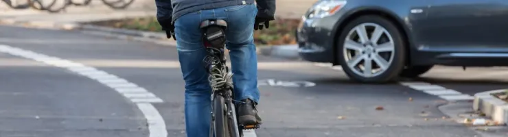 Un cycliste vu de dos roule sur une piste cyclable, avec une voiture grise garée à côté.