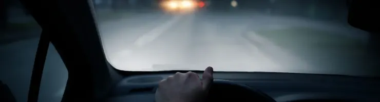 Une main tient le volant d'une voiture, les phares d'un autre véhicule sont visibles de nuit à travers le pare-brise.
