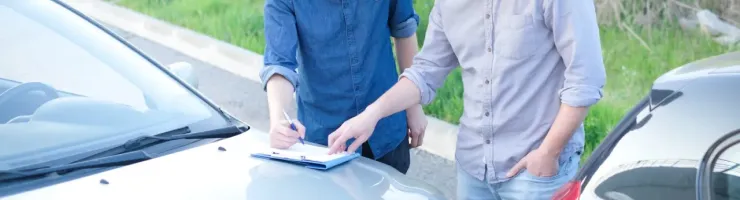 Deux hommes sont penchés sur le capot d'une voiture endommagée, l'un d'eux remplit un constat à l'amiable sur un presse-papiers.