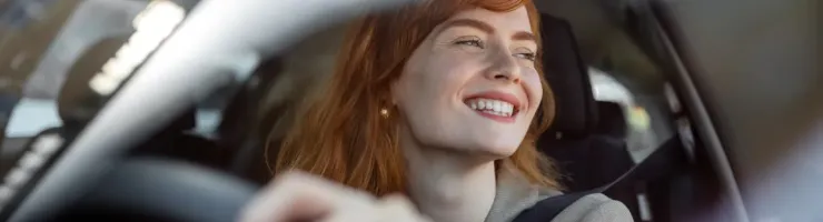 Une femme rousse souriante et joyeuse est vue à travers le pare-brise, conduisant sa voiture.