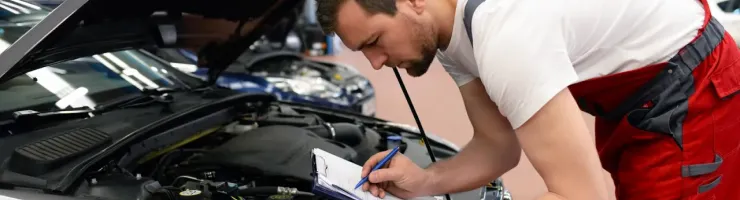Un mécanicien en salopette rouge et blanche est penché sur le moteur d'une voiture, notant des informations sur un presse-papiers.