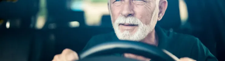 Un homme âgé aux cheveux et à la barbe blancs conduit une voiture, le regard concentré.