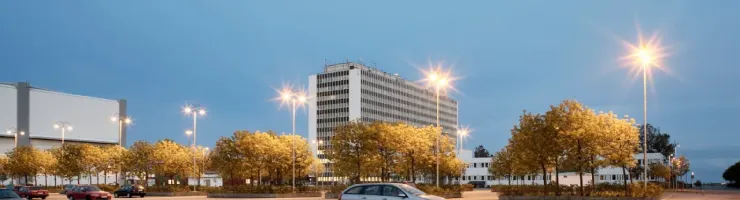 Un parking presque vide, une voiture grise est au milieu de l'iamge et un bâtiment moderne en arrière-plan, éclairé par des lampadaires.