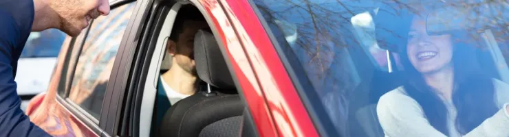 Un homme souriant se penche vers une femme elle aussi souriante assise au volant d'une voiture rouge.