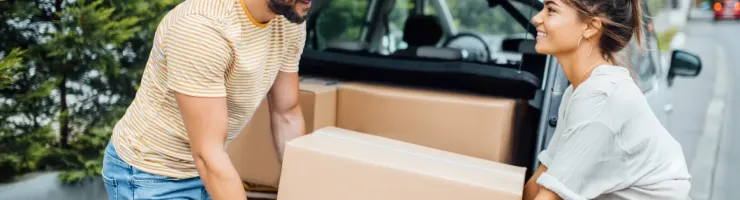 Un jeune couple charge des cartons dans le coffre ouvert d'une voiture, souriant l'un à l'autre.