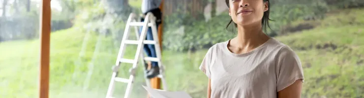 Une femme tenant un plan devant une fenêtre et un homme sur un escabeau qui prend des mesures en arrière-plan.