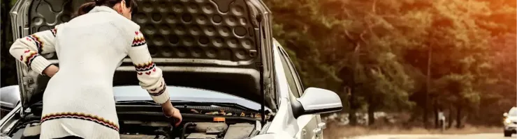 Une femme regarde sous le capot ouvert d'une voiture argentée en panne sur le bord de la route.