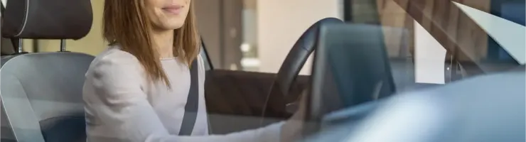 Une femme, souriante, est assise au volant d'une voiture, attachée par sa ceinture de sécurité, en train d'utiliser l'écran tactile de sa voiture.