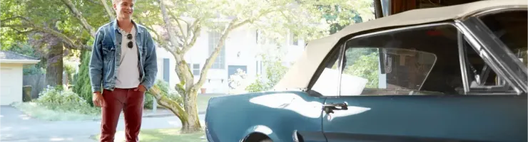 Un jeune homme aux cheveux blonds clairs se tient à côté d'une voiture décapotable bleue vintage dans un garage.