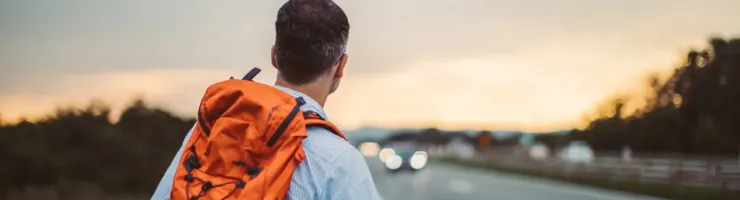 Un homme avec un sac à dos orange fait de l'auto-stop au bord d'une route au coucher du soleil, dos à la caméra.