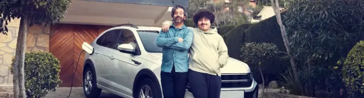 Un père et son fils souriants posent devant une voiture électrique blanche branchée à une borne de recharge devant une maison.