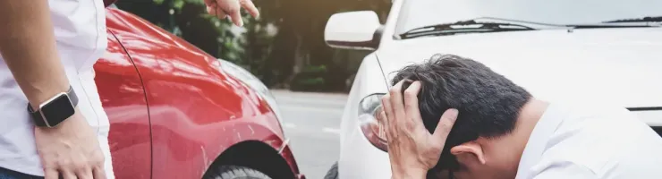 Un homme se tient la tête, désespéré, après un accident de voiture, tandis qu'une autre personne pointe du doigt la voiture rouge.