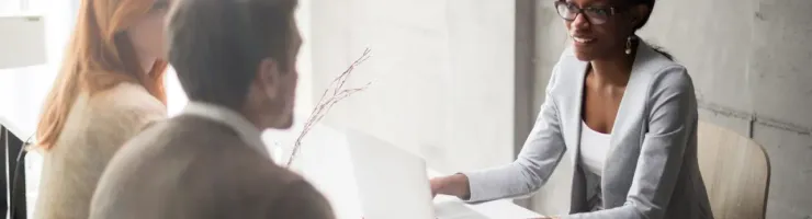Une conseillère souriante en lunettes discute avec un couple assis à son bureau devant un ordinateur portable.