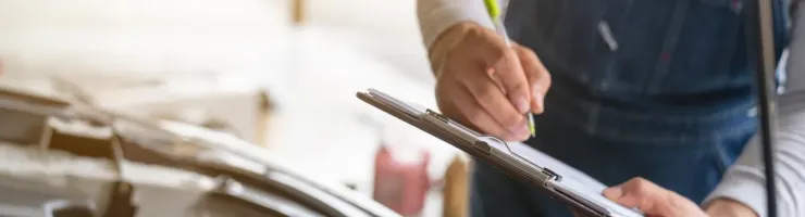 Un mécanicien en salopette écrit sur un presse-papiers, effectuant une inspection sous le capot d'une voiture.