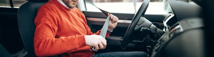 Un homme en pull orange et lunettes attache sa ceinture de sécurité dans une voiture, se préparant à conduire.