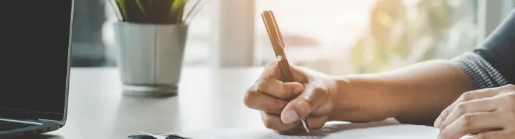 Une personne signe un document avec un stylo sur un bureau, à côté d'un ordinateur portable et d'une plante verte.
