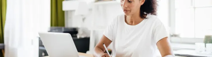 Une femme assise à domicile, écrivant sur un papier et regardant son ordinateur portable.