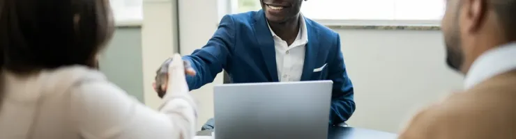 Un homme souriant en costume bleu serre la main d'une femme à un bureau, avec un ordinateur portable ouvert.