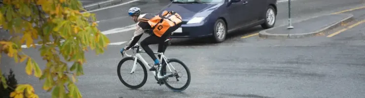 Un cycliste avec un grand sac à dos orange roule sur une route mouillée, avec une voiture en arrière-plan.