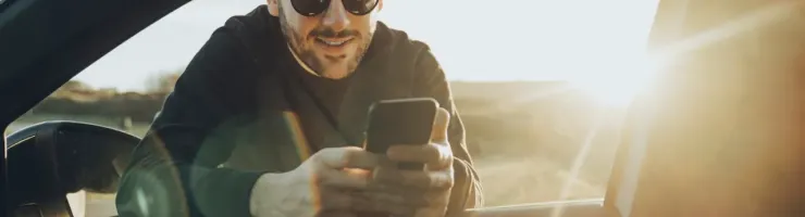 Un homme souriant et portant des lunettes de soleil regarde son téléphone à travers la fenêtre ouverte d'une voiture.