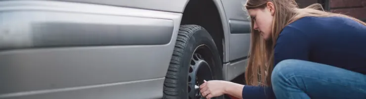 Une femme est accroupie à côté d'une voiture grise, en train de changer une roue ou de serrer les écrous.