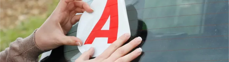 Des mains appliquent un autocollant "A" rouge pour jeune conducteur sur la lunette arrière d'une voiture.