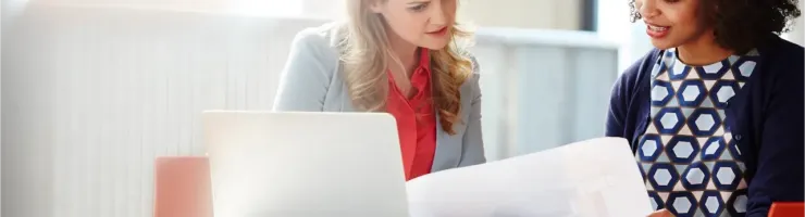 Deux femmes discutent et examinent des documents devant un ordinateur portable sur une table.