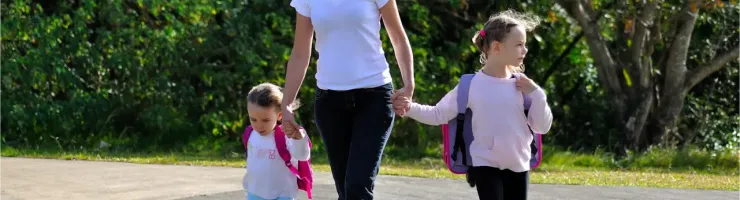 Une femme tient la main de deux jeunes enfants avec des sacs à dos, traversant la route sur un passage clouté.