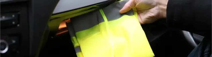 Une main retire un gilet de sécurité jaune fluorescent du vide-poche d'une voiture.
