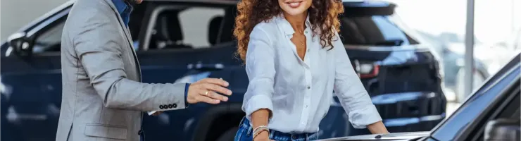 Un vendeur sourit à une femme aux cheveux bouclés qui regarde le capot d'une voiture.