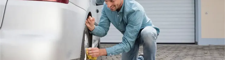 Un homme souriant nettoie la roue arrière d'une voiture grise devant un garage.