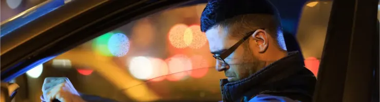 Un homme l'air fatigué au volant d'une voiture la nuit, avec des lumières urbaines floues.