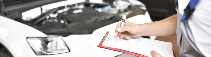 Un mécanicien en salopette prend des notes sur un presse-papiers devant un moteur de voiture ouvert.