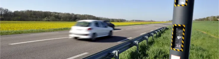 Un radar automatique noir au bord d'une route avec une voiture blanche en mouvement.