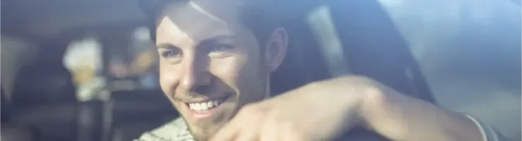 Un jeune homme souriant au volant d'une voiture, regardant à travers le pare-brise.
