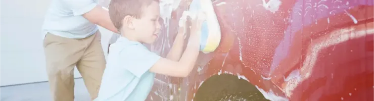 Un jeune garçon savonne une voiture rouge avec une éponge, aidé par un adulte en arrière-plan.